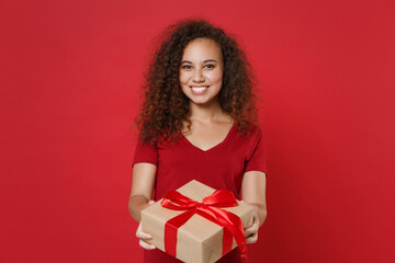 Smiling young african american woman girl in casual t-shirt posing isolated on red wall background. St. Valentine's Day, Women's Day, birthday, holiday concept. Hold present box with gift ribbon bow.