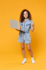 Full length portrait of smiling young african american woman girl in denim clothes isolated on yellow background in studio. People lifestyle concept. Working on laptop pc computer, showing thumb up.
