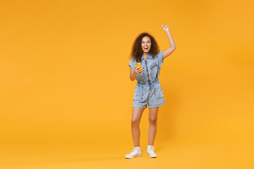 Fototapeta na wymiar Full length portrait of happy young african american woman girl in denim clothes isolated on yellow wall background in studio. People lifestyle concept. Using mobile cell phone, doing winner gesture.
