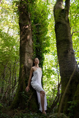 junge Frau in weißem Kleid an Baum gelehnt genießt die Atmosphäre im Wald, wahrnehmen, Verbindung und neuendecken von Natur.