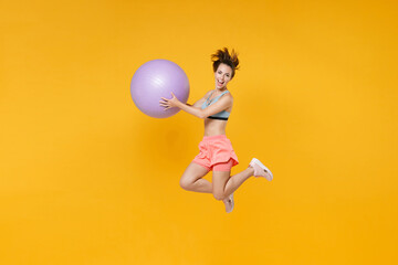Fototapeta premium Full length portrait cheerful young fitness woman in sportswear working out isolated on yellow background studio. Workout sport motivation lifestyle concept. Mock up copy space. Jumping, hold fitball.