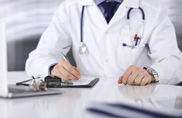 Unknown male doctor sitting and working with clipboard of medication history record in clinic at his working place, close-up. Young physician at work. Perfect medical service, medicine concept