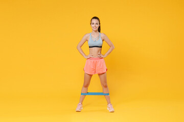 Full length portrait of smiling young fitness woman girl in sportswear working out isolated on yellow background. Workout sport motivation lifestyle concept. Doing exercise for legs with fitness gums.