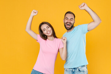 Cheerful young couple friends guy girl in blue pink t-shirts posing isolated on yellow wall background studio. People lifestyle concept. Mock up copy space. Keeping eyes closed, rising hands, dancing.