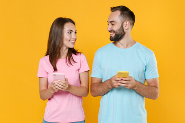 Smiling young couple friends guy girl in blue pink t-shirts isolated on yellow background. People lifestyle concept. Mock up copy space. Using mobile phones typing sms messages, looking at each other.