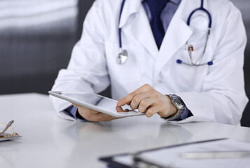 Unknown male doctor sitting and working with tablet computer in clinic at his working place, close-up. Young physician at work. Perfect medical service, medicine concept