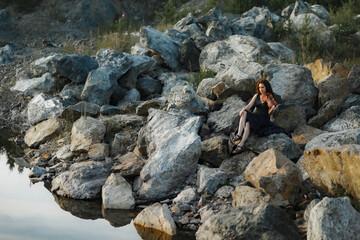 brunette girl with lush curly hair sits on the stones near the lake in summer in a dark blue dress with a violin, professional musician violinist