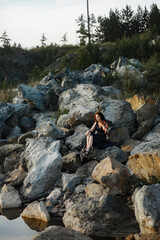 brunette girl with lush curly hair sits on the stones near the lake in summer in a dark blue dress with a violin, professional musician violinist