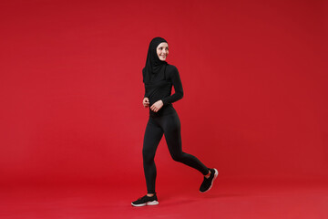 Fototapeta na wymiar Full length portrait of smiling young arabian muslim woman in hijab black clothes posing isolated on red background. People religious lifestyle concept. Mock up copy space. Running, looking aside.