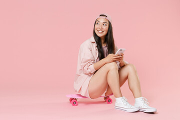 Obraz na płótnie Canvas Full length portrait of smiling young asian woman girl in casual clothes, cap isolated on pastel pink background . People lifestyle concept. Sit on skateboard, using mobile cell phone, looking aside.