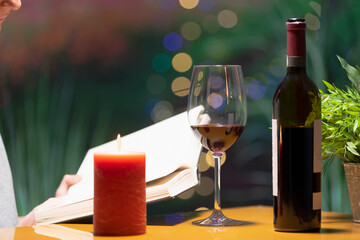 a glass of wine surrounded by a lit candle and a bottle of wine, with an open book being held by a person