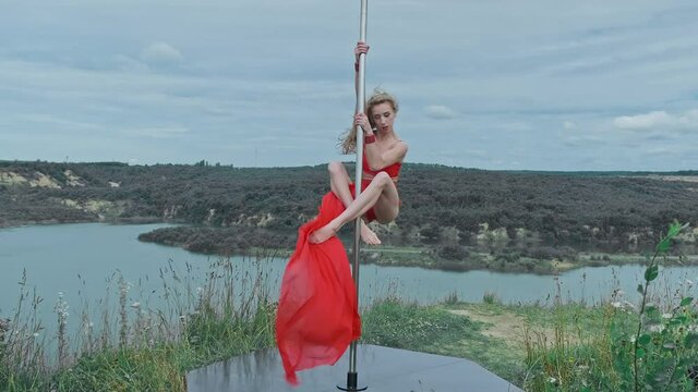 Young woman in red dancing on pole. Female dancer pole dancing outdoors. Sky and lake at background. 4K, UHD