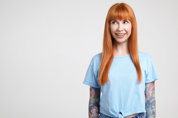 Cheerful young attractive long haired redhead lady with casual hairstyle smiling widely while looking gladly aside, posing over white background in blue t-shirt