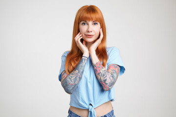 Charming young green-eyed long haired tattooed lady smiling gently while looking positively at camera and keeping raised hands on her face, isolated over white background