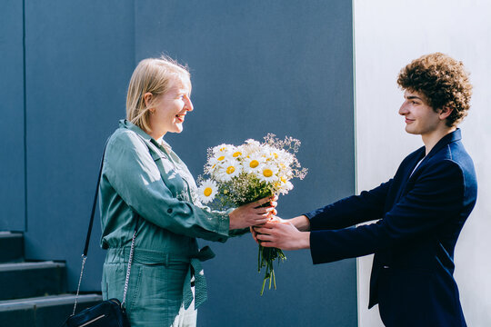 Young Curly Man Gives Flowers Older Woman. Smiling Adult Son Giving Flowers Daisies Bouquet To His Senior Mother Outdoor.