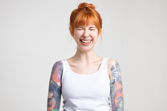 Joyful young attractive redhead woman with tattoos keeping her eyes closed while laughing cheerfully, dressed in white shirt while posing over white background