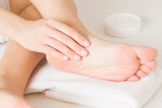 Young woman hand applying moisturizing cream on foot. Barefoot on white towel. Jar with natural herbal cream. Care about clean and soft legs skin. Healthcare concept. Closeup.