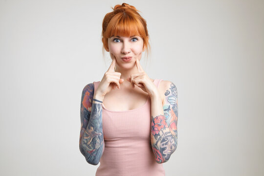 Studio photo of attractive redhead female with tattoos keeping index fingers on her cheeks while fooling, standing over white background in nude shirt