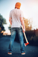 A young man in a fashionable yellow hat and blue jeans stands confidently with a sharp large axe in...