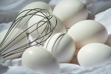 White chicken eggs and a whisk on a white cloth.