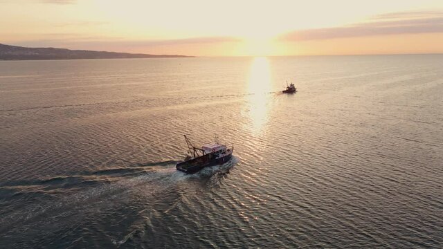 Two fishing boats sail at sunrise to catch school of fish on calm sea surface in summer. Commercial catch of sea fish. Small ship floats on sea surface leaving path of sea foam water. Aerial view