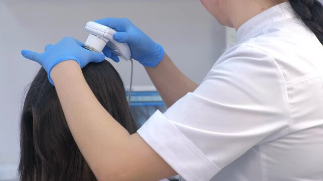 Doctor trichologist examines woman patient's hairs using trichoscope in clinic, closeup hand view. Runs a trichoscope over scalp and sees result on monitor screen. Procedure of computer trichoscopy.
