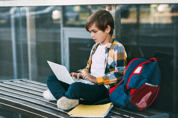 A cute boy in a plaid shirt is sitting on a bench with a laptop and typing on the keyboard, next to a backpack. The student is preparing for outdoor classes