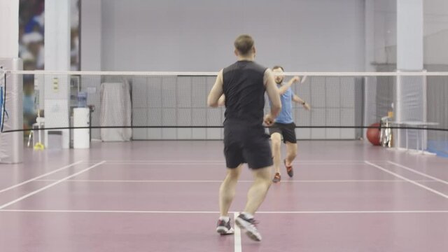 Sportive Caucasian men playing badminton indoors. Wide shot portrait of two young sportsmen exercising in gym. Athletic guys training on indoor court with rackets and shuttlecock.