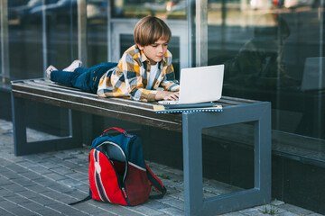 A schoolboy is lying on a wooden bench and working on a laptop, next to a backpack. The boy is preparing for school lessons using the Internet. Social distance. Distance learning,
