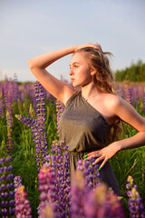 A blonde girl in an elegant grey dress with an open shoulder poses in lupine fields.