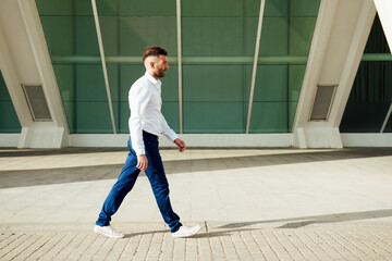 young man in white shirt, blue pants and short beard walking sideways to camera