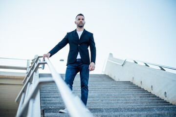 Casual stylish young man in jacket and jeans walking down staircase with hand on palm rest