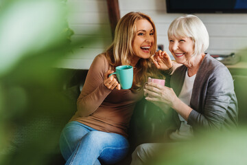 Happy senior mother with adult daughter sitting on couch and holding cups with coffee or tea at...
