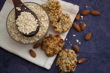 Oatmeal in a jar and wooden spoon on white serviette with oatmeal cookies and almond around it. Violet grunge background
