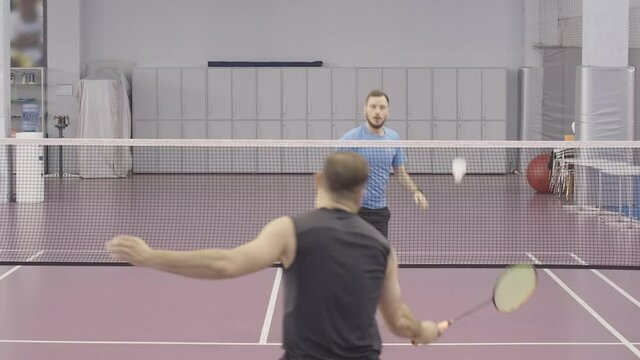 Young athletic Caucasian sportsman in blue T-shirt playing badminton with brunette man in black sportswear. Portrait of confident sportive men training in gym on indoor court.