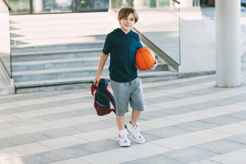 Fototapeta premium A cute boy with a backpack in one hand and a basketball in the other. The boy in shape in a hurry for a workout but basketball. Training, education, physical education