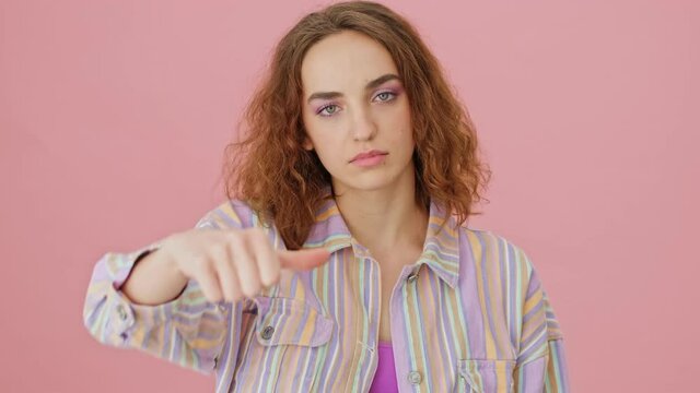 A serious young woman is showing thumb down gesture to the camera standing isolated over a pink background in studio