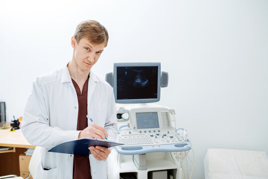 Portrait of a handsome young doctor next to an ultrasound scanning machine. He's squinting, looking at the camera with untrusting look, filling patient's history.