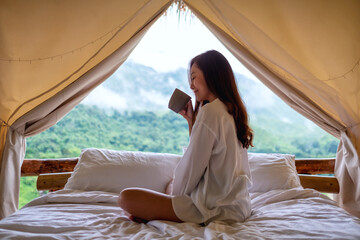 Portrait image of a beautiful young asian woman drinking coffee while sitting on a white bed in the morning with a beautiful nature view outside the tent