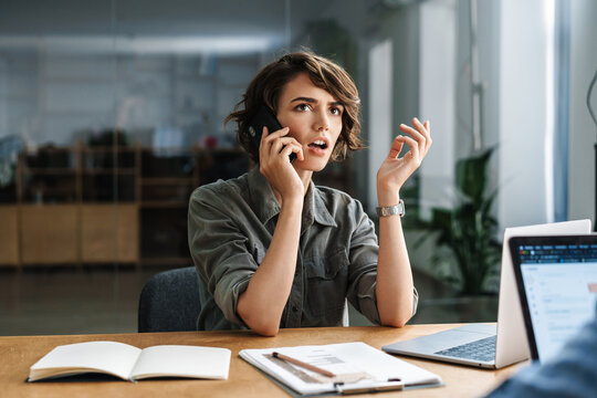 Image of displeased woman talking on cellphone while working with laptop