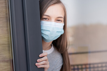 Concept of coronavirus quarantine. Child wearing medical protective face mask during flu virus, looking out of window. COVID-19 - self isolation. Teen girl forced to stay at home. View from street.