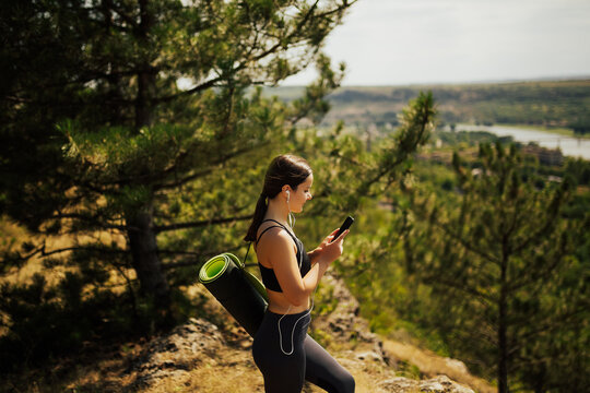 Cute smiling woman using smartphone and holding yoga mat on the hill. Pretty young carrying a yoga mat and texting on her mobile phone outdoors.