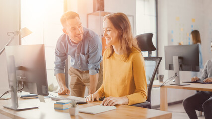 Female Specialist Works on Desktop Computer, Project Manager Stands Beside and gives Advice on Optimizing Workflow for Customer Experience Management. Shot with Warm Sun Flare in Modern Office