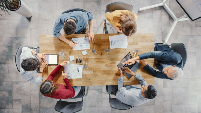 Startup Meeting Room: Team Of Entrepreneurs Sitting At The Conference Table Have Discussions, Solve Problems, Use Digital Tablet, Laptop, Share Documents With Statistics, Charts. Top View