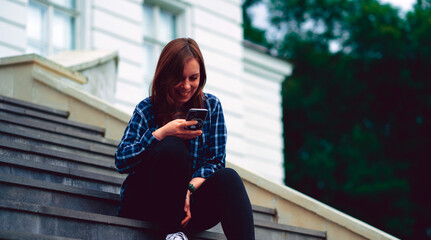 Young woman flipping news in smartphone, sitting on stairs of manor. Adult female with mobile phone sitting on steps of Palace.