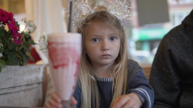 A funny little girl sits in a cafe with a large glass of strawberry milk cocktail. 