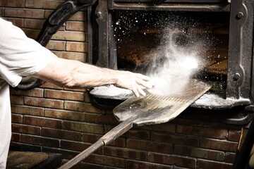 Beautiful shot of a baker pouring flour on the surface of a peel near a masonry brick oven