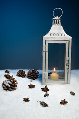 White Christmas lantern on snow with pine cones, cinnamon stars in front of dark blue background, copy space