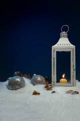 White Christmas lantern on snow with pine cones and big copper baubles in front of dark blue background, copy space