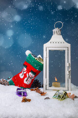 White Christmas lantern on snow with pine cones and colorful christmas presents in the snow and in a stocking with snowman motive in front of dark blue background